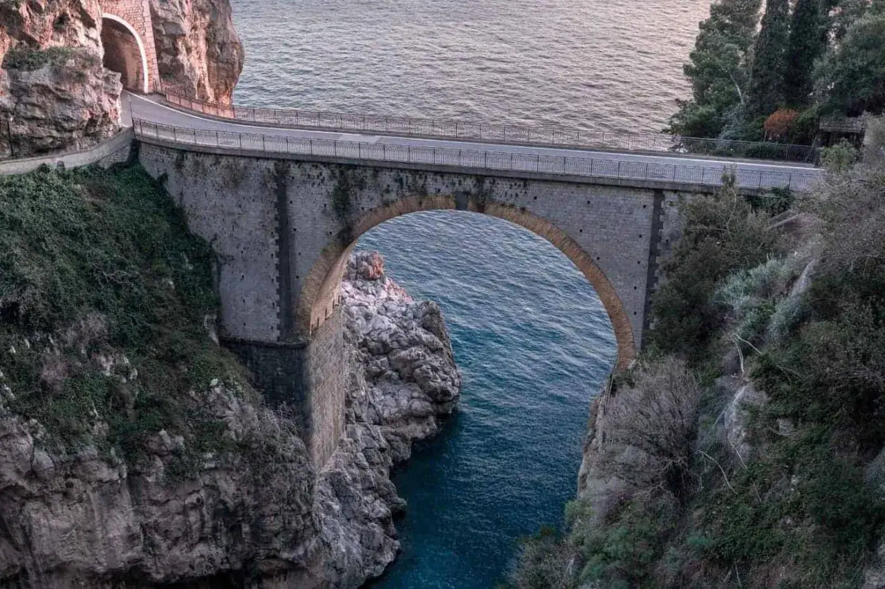 Ponte ad arco in pietra che attraversa un fiordo, con il mare blu sottostante. Un'icona del furore costiera amalfitana, incastonata tra rocce e vegetazione.