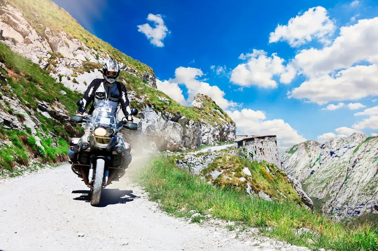 Motociclista affronta un ripido tornante della boaria su una strada sterrata in montagna, con un cielo azzurro e nuvole bianche.