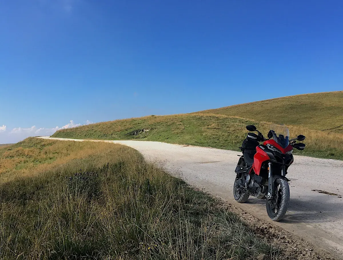 Ducati rossa ferma su strada sterrata, con un passo branchetto sullo sfondo e cielo azzurro.