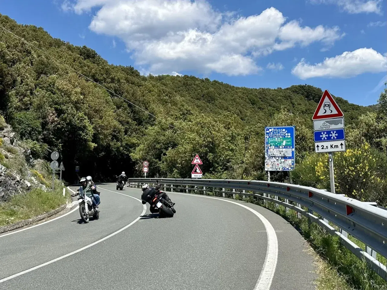 Motociclisti in curva sul passo del Bracco, tra il verde della Liguria e un cielo azzurro con nuvole bianche.