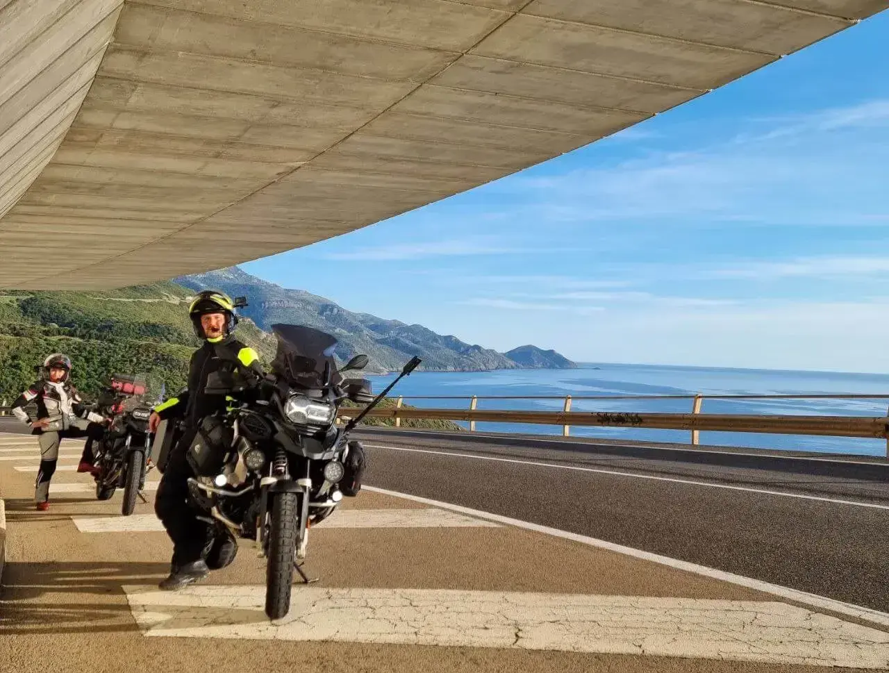 Motociclisti in sosta panoramica, con moto pronte per esplorare la costa tra Alghero e Oristano. Mare azzurro e montagne sullo sfondo.