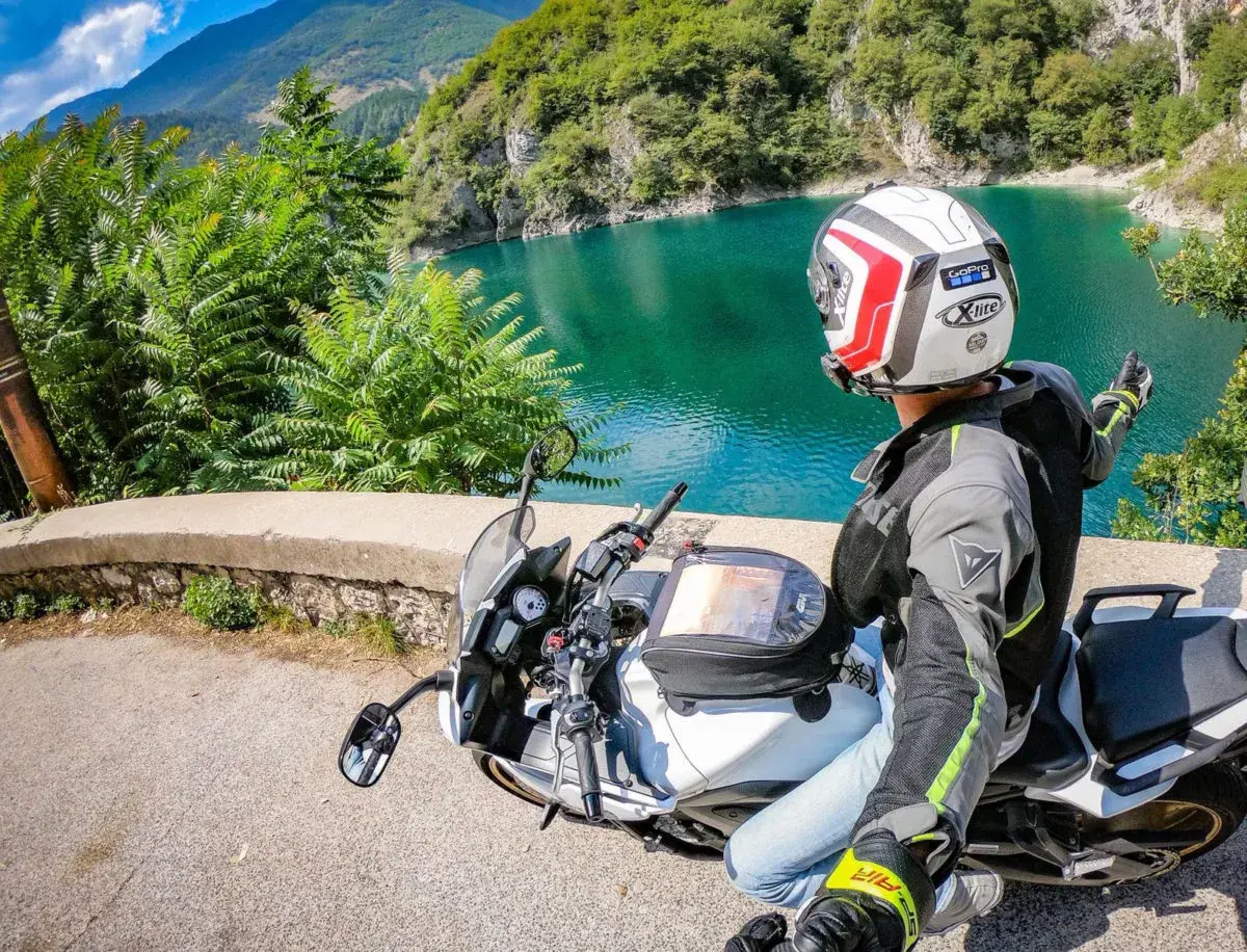 Weekend in moto nel Nord Italia: un motociclista ammira un lago alpino turchese, con montagne verdi sullo sfondo.