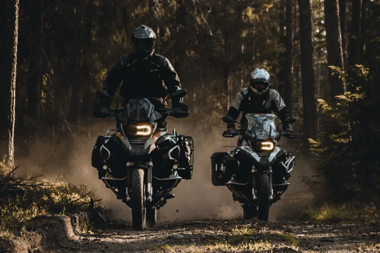 Due motociclisti avventurosi sfrecciano su una strada sterrata nel bosco, sollevando polvere.