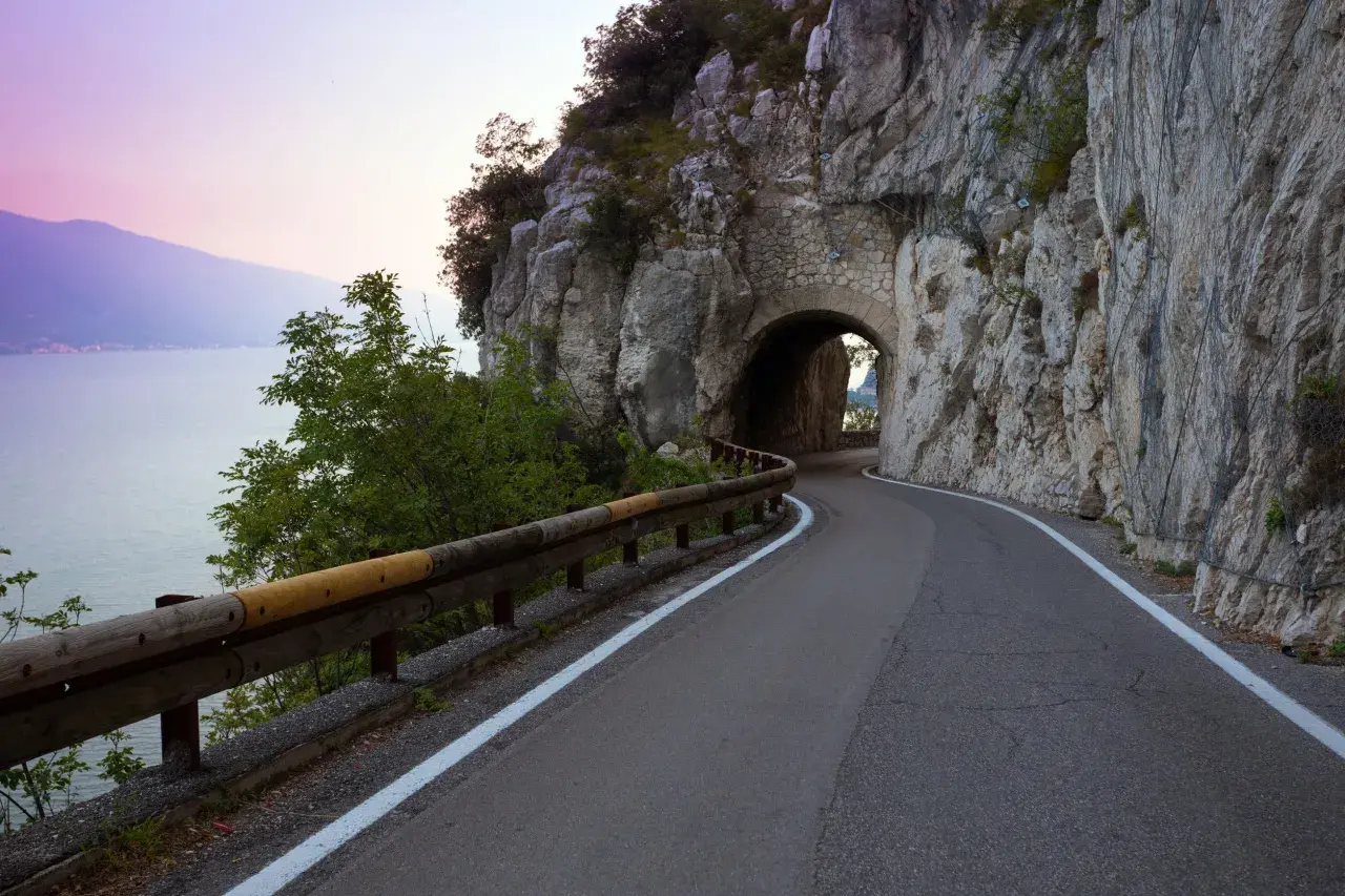 Strada della forra in moto, con un tunnel scavato nella roccia e il lago sullo sfondo al tramonto.