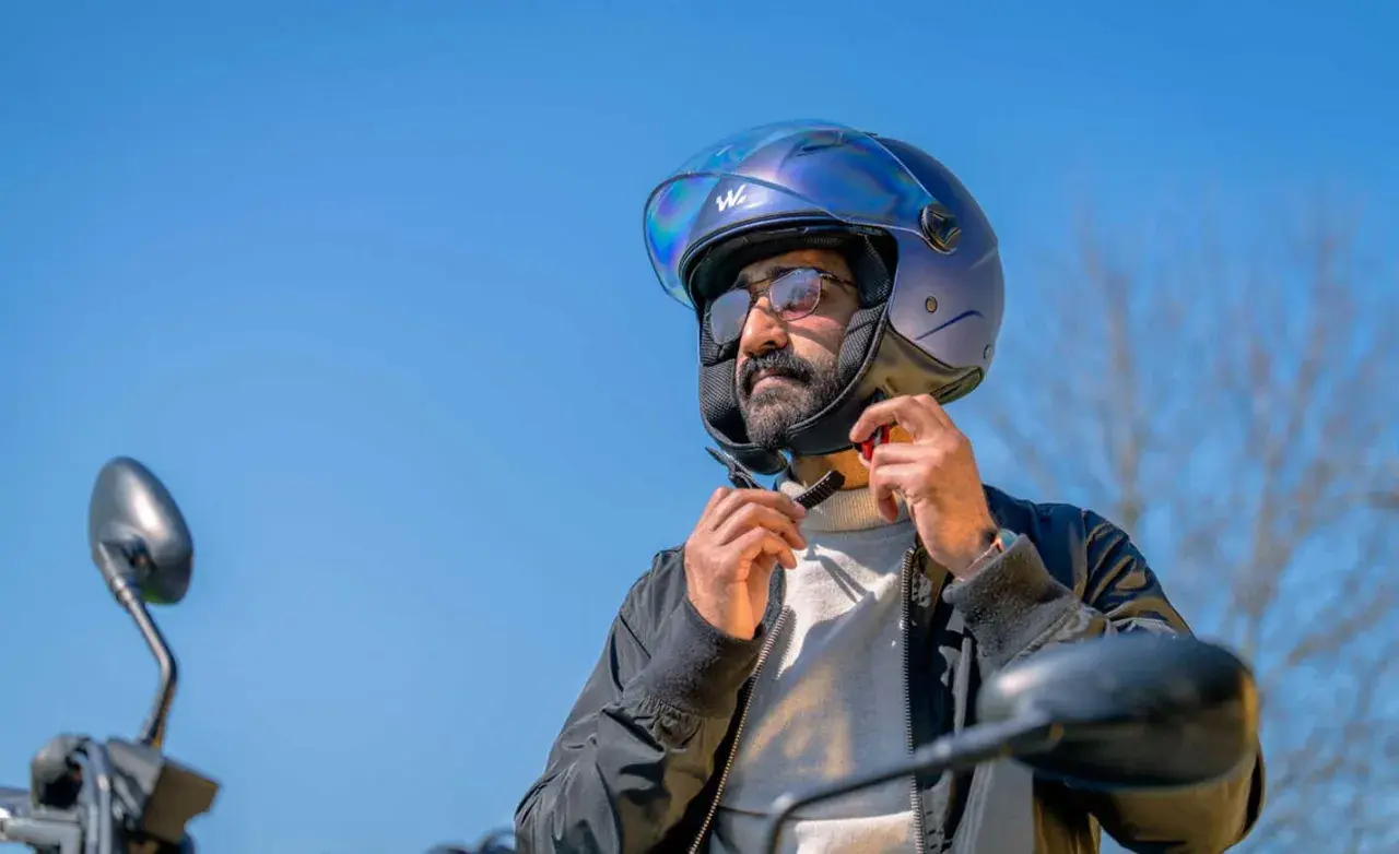 Uomo con casco moto blu, occhiali e barba, che allaccia il sottogola. Ideale per chi cerca una tabella misure casco moto.