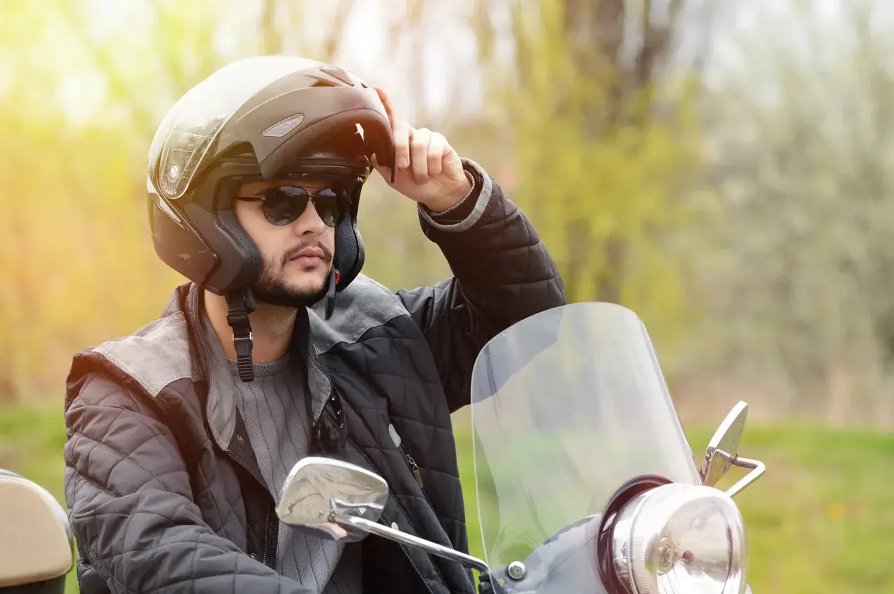 Uomo con casco da moto, occhiali da sole e giacca imbottita, che regola le parti del casco.