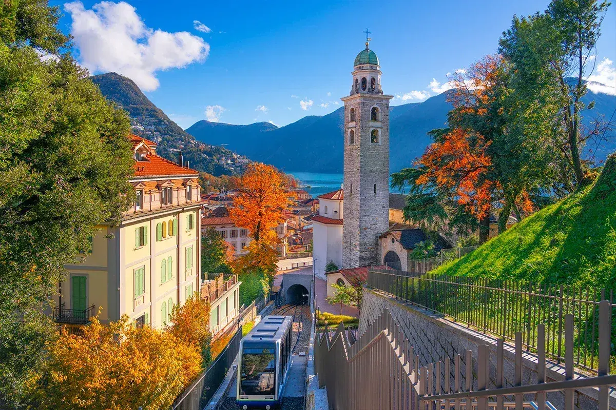 Un trenino scende tra case colorate e un campanile, con il Lago Maggiore sullo sfondo. Un assaggio delle **strade panoramiche Lago Maggiore** in autunno.