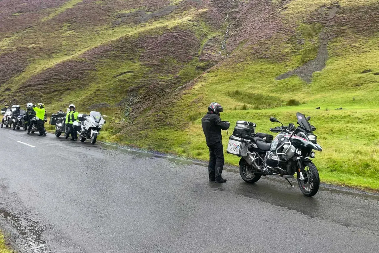 Motociclisti in sosta sul passo della Contessa, con moto cariche e un paesaggio collinare verde e viola sullo sfondo.
