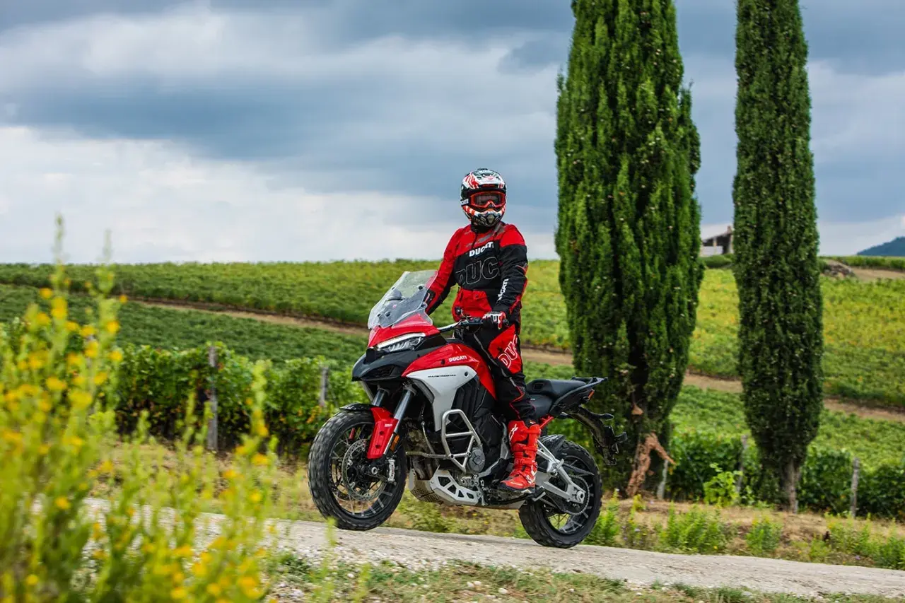 Un motociclista in tuta rossa sfreccia tra i cipressi e i vigneti del Val d'Orcia in moto.