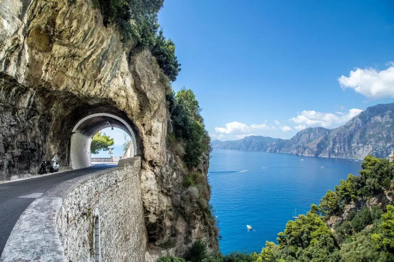 Strada panoramica a picco sul mare blu intenso, con un tunnel scavato nella roccia e una vista mozzafiato su Meta di Sorrento.