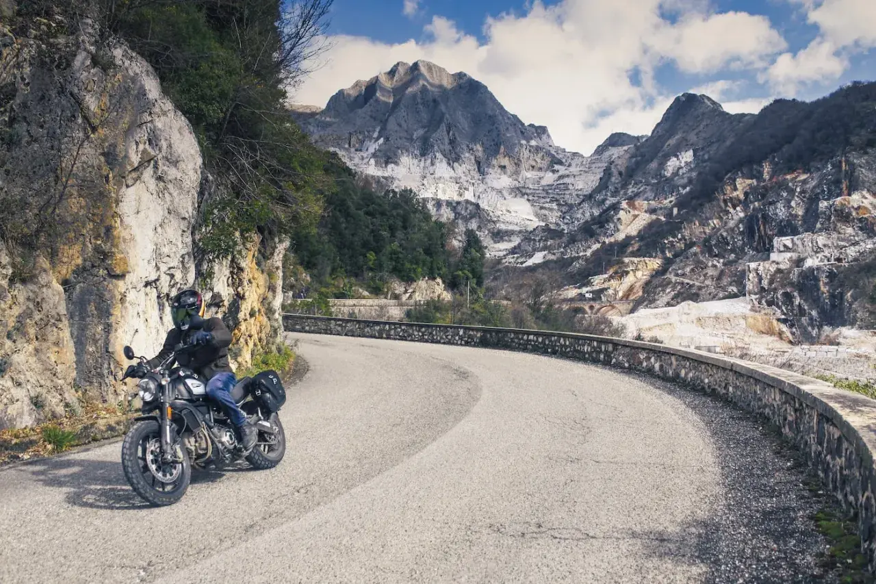 Motociclista su strada panoramica toscana, con montagne di marmo sullo sfondo.