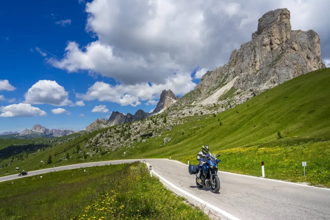 Motociclista percorre la strada cimina tra prati verdi e montagne imponenti sotto un cielo azzurro con nuvole bianche.