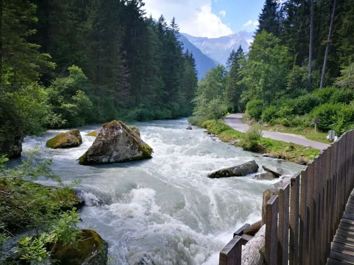 Fiume impetuoso tra rocce e boschi, con una strada serpeggiante e montagne sullo sfondo, ricorda le cascate del Nardis.