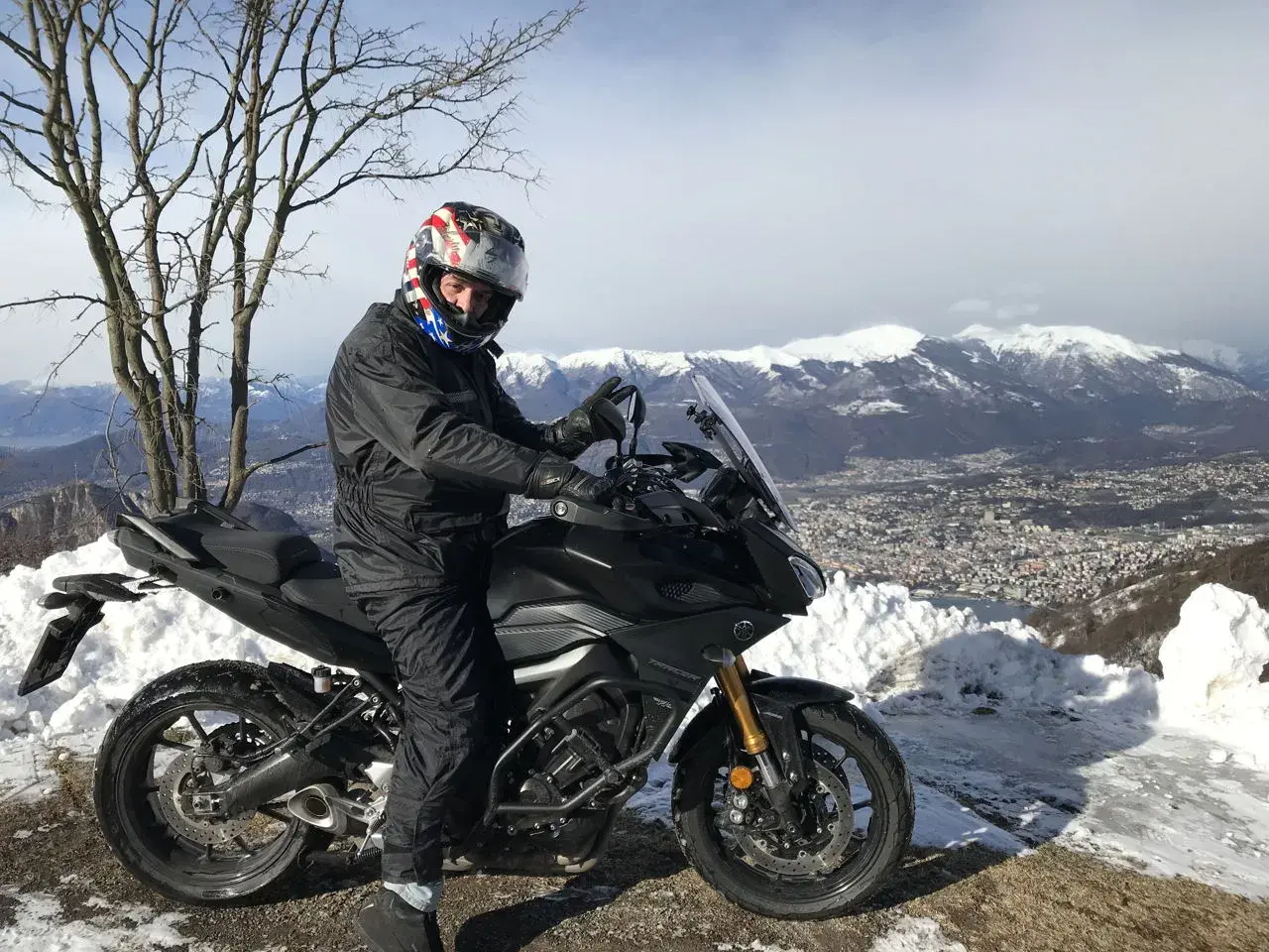 Motociclista con casco stellato su un balcone d'Italia innevato, con vista su montagne e città.