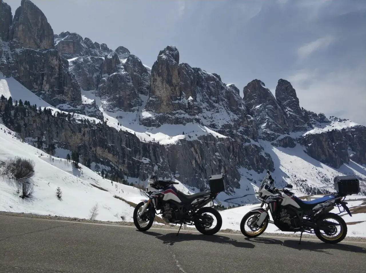 Due moto da enduro parcheggiate su una strada innevata, con imponenti vette delle valli bergamasche sullo sfondo.