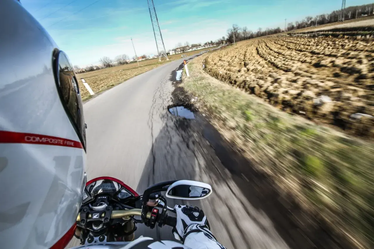 In sella a una moto, con il casco bianco e rosso, si percorre una strada di campagna. Il campo arato a lato e il cielo azzurro evocano il linguaggio motociclisti italiano.