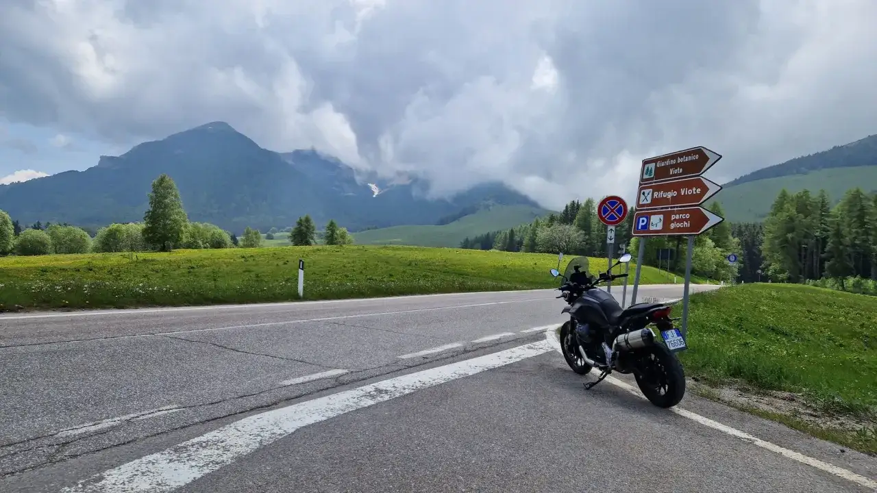 Moto ferma su strada panoramica, con montagne nebbiose e prati verdi sullo sfondo. Un cartello indica 