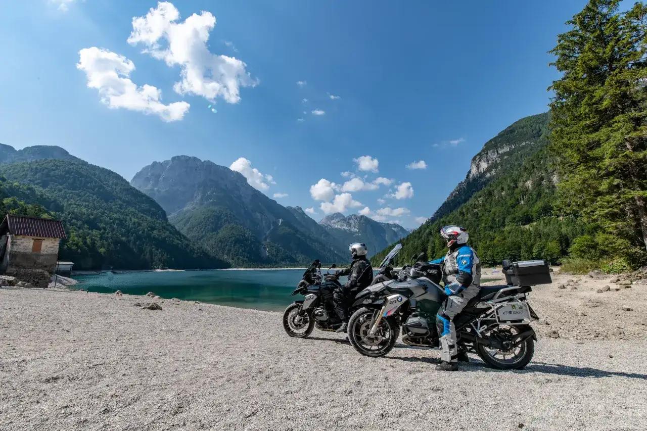 Due motociclisti si fermano con le loro moto su una spiaggia ghiaiosa, con un lago turchese e montagne imponenti sullo sfondo, un paesaggio mozzafiato al passo del Tarvisio.