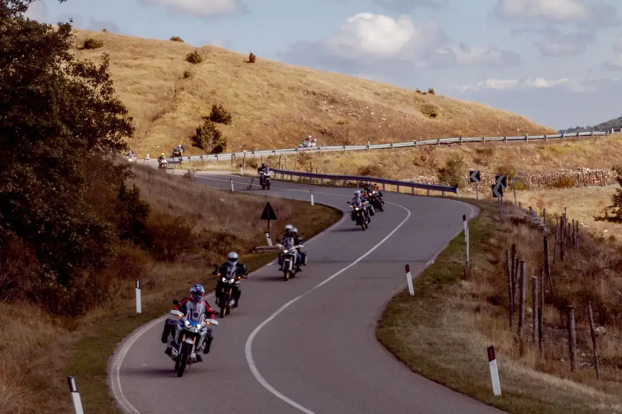 A group of motorcyclists ride along a winding mountain road with dry, grassy hillsides.
