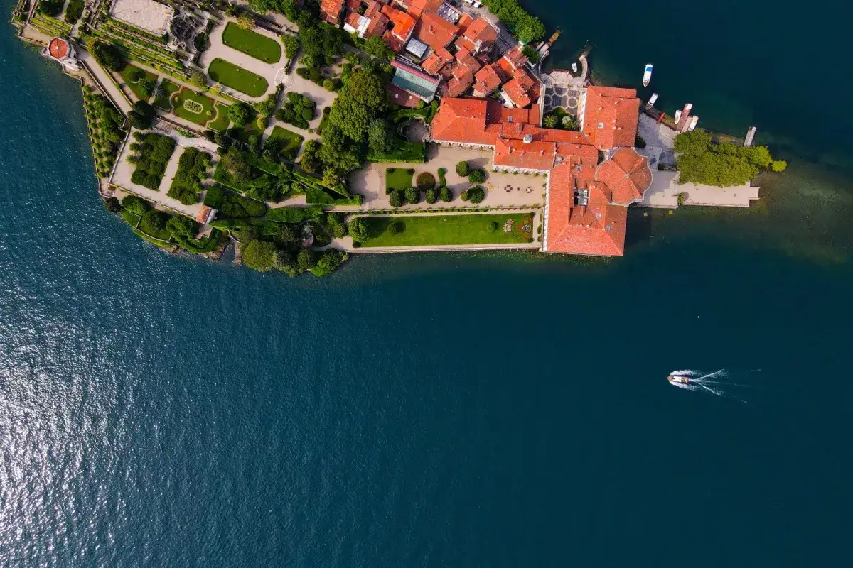 Vista aerea di un'isola con giardini curati e palazzi dai tetti rossi, mentre una barca solca il blu intenso del lago. Un'idea per un giro del Lago Maggiore in moto.