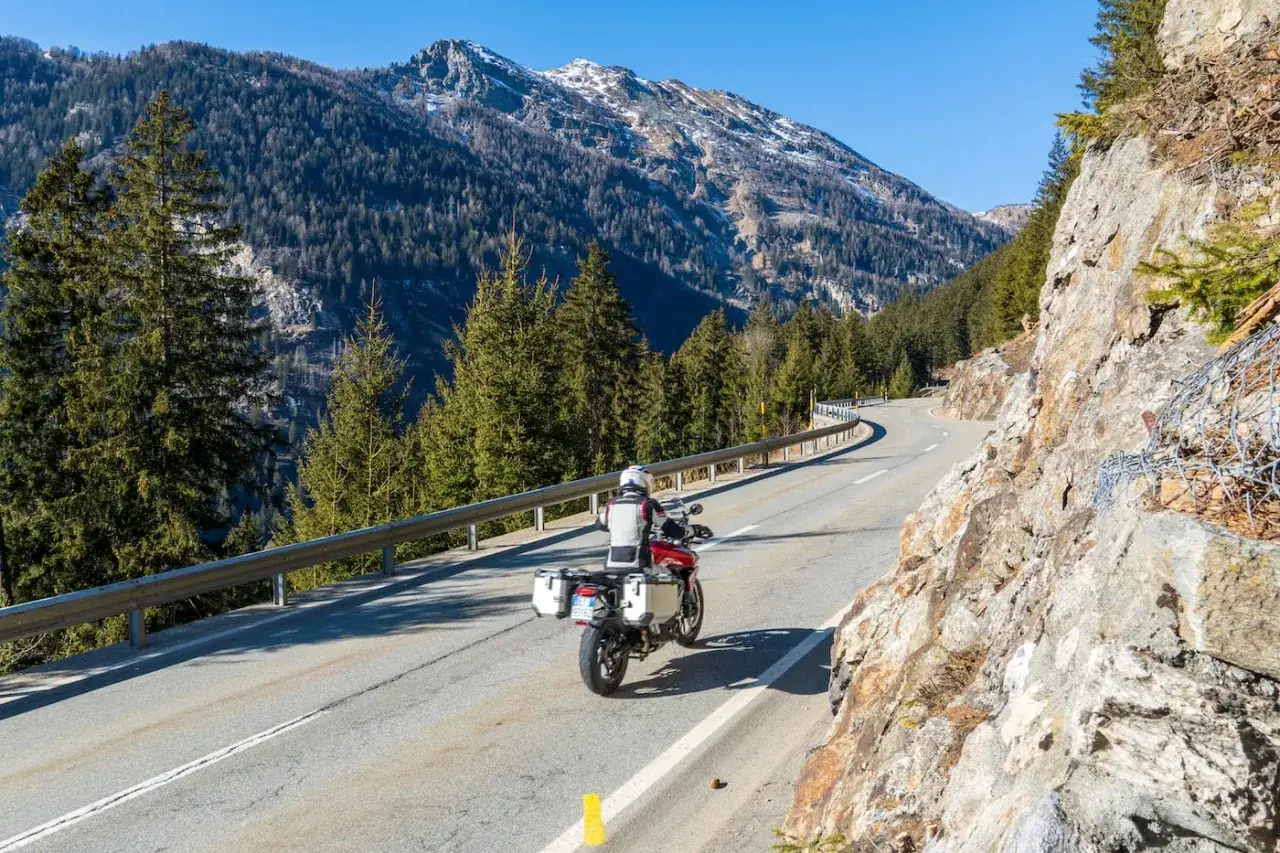 Un motociclista percorre una strada tortuosa in Valtellina, circondato da imponenti montagne e fitti boschi di conifere.