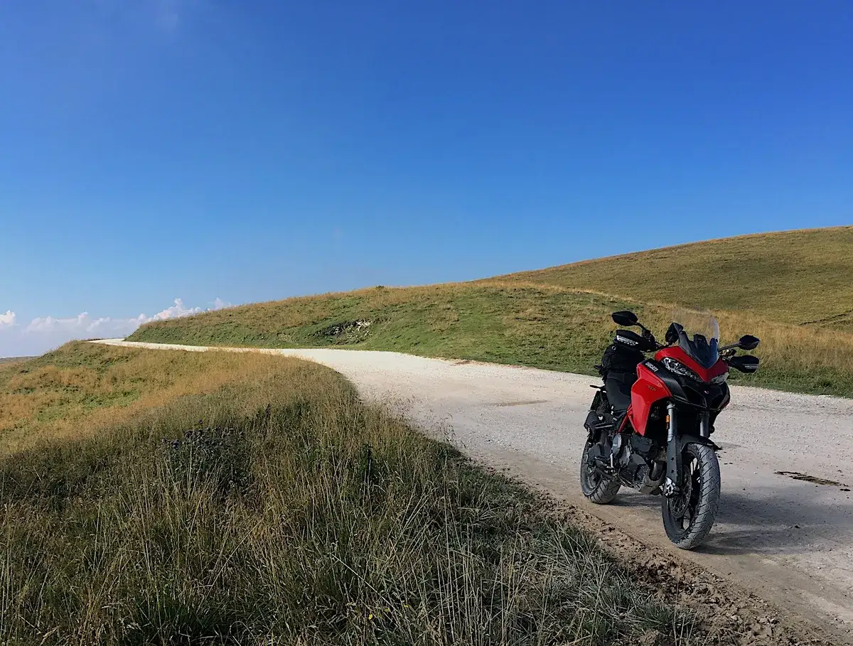 Ducati rossa ferma su strada sterrata, con un passo branchetto sullo sfondo e cielo azzurro.