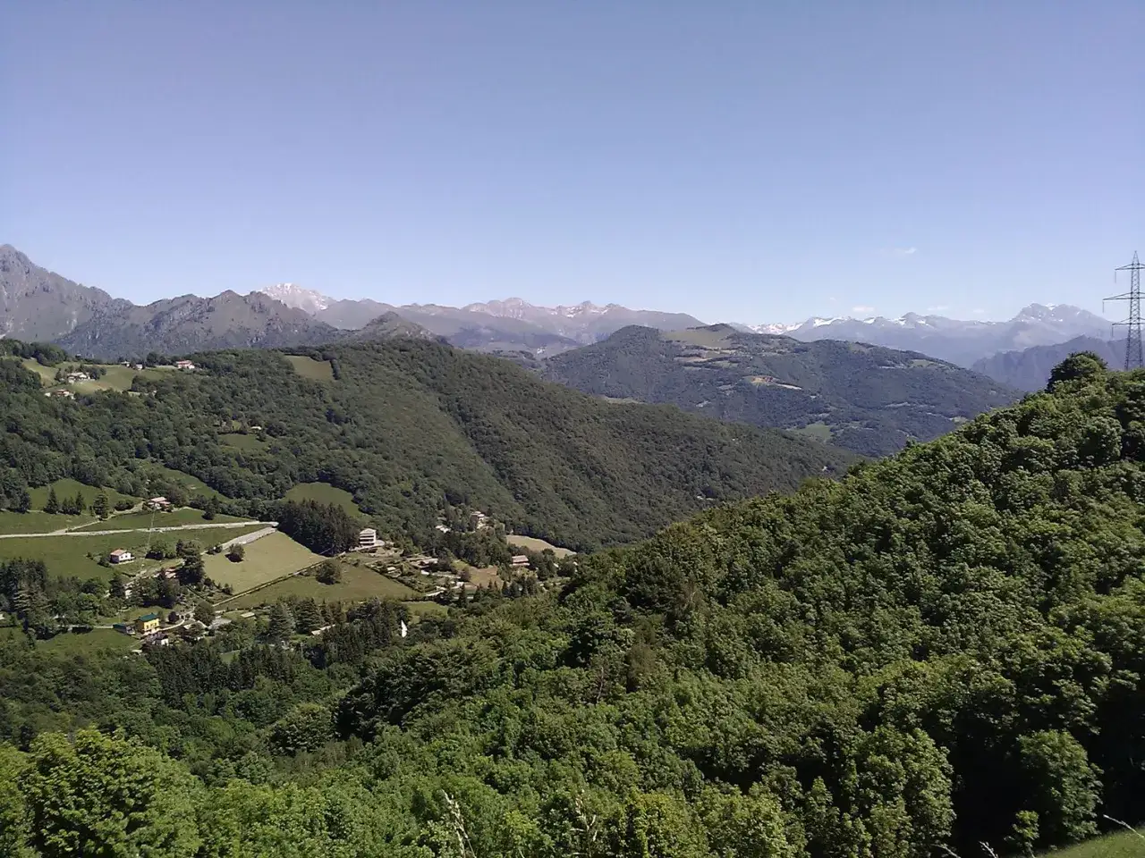Panoramica mozzafiato dal passo di Ganda, con dolci colline verdi, case sparse e montagne innevate all'orizzonte.