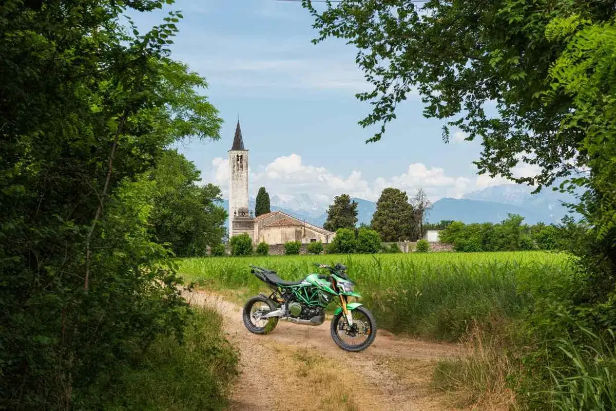 Moto verde su sterrato, con chiesa e montagne sullo sfondo, in un paesaggio tipico dei passi del Friuli Venezia Giulia.