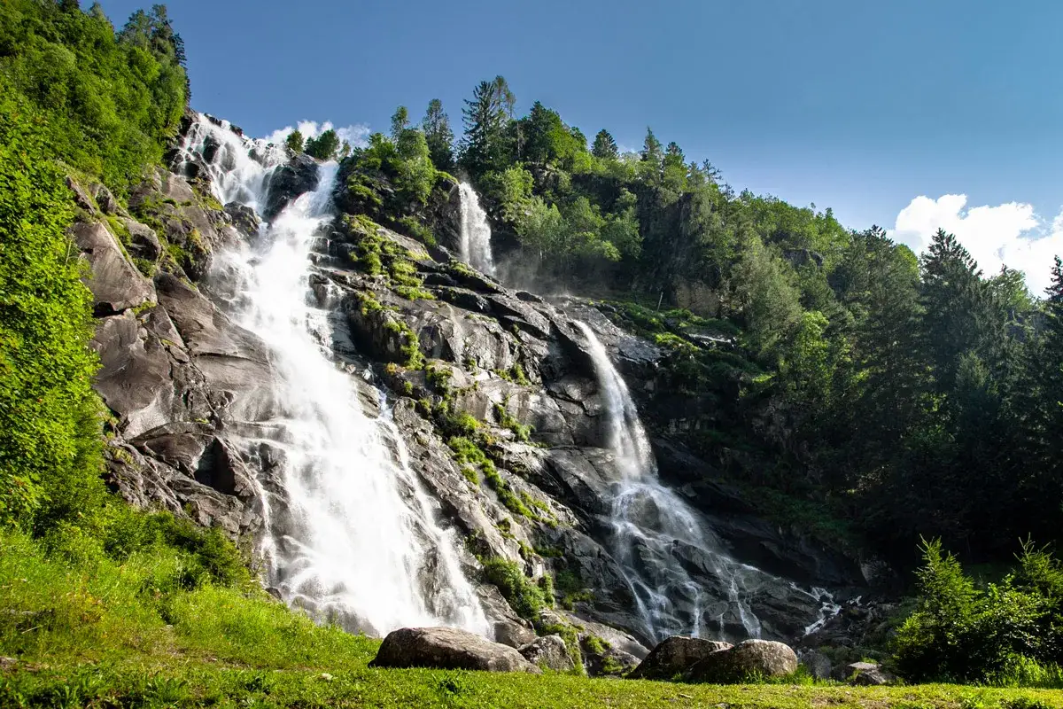 Le maestose cascate del Nardis scendono impetuose su rocce scure, immerse nel verde lussureggiante di una foresta alpina sotto un cielo azzurro.