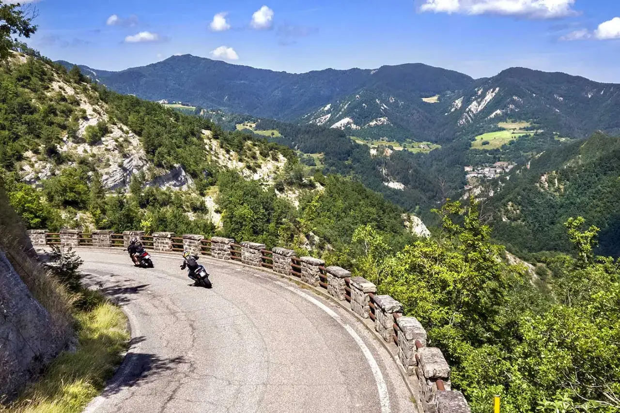Due motociclisti affrontano una curva panoramica sulla strada statale 67, tra montagne verdi e un cielo azzurro.