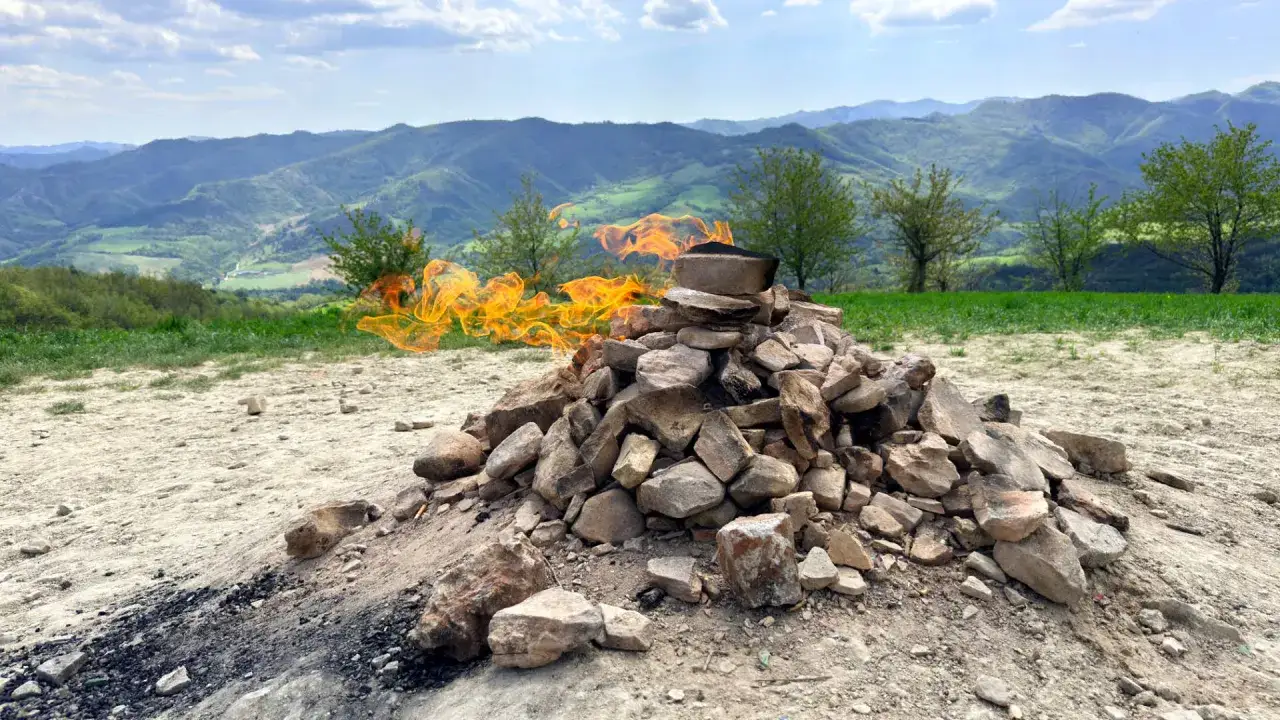 Fiamme danzano su un cumulo di pietre in cima al monte Busca, con dolci colline verdi sullo sfondo.