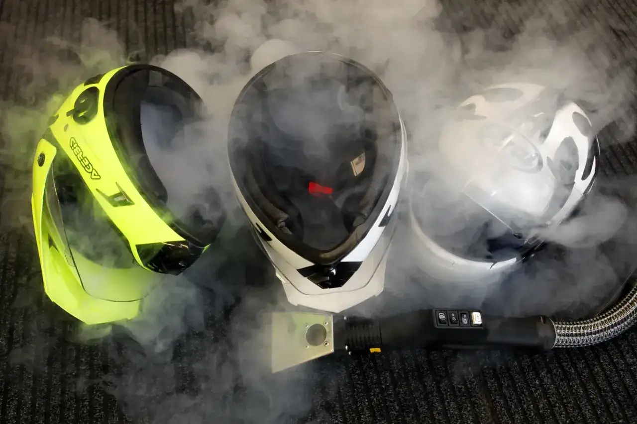 Tre caschi, uno giallo fluo, uno bianco e nero, uno bianco, circondati da vapore. Un pulitore a vapore &egrave; posizionato davanti, suggerendo che il casco non deve essere pulito con solventi.