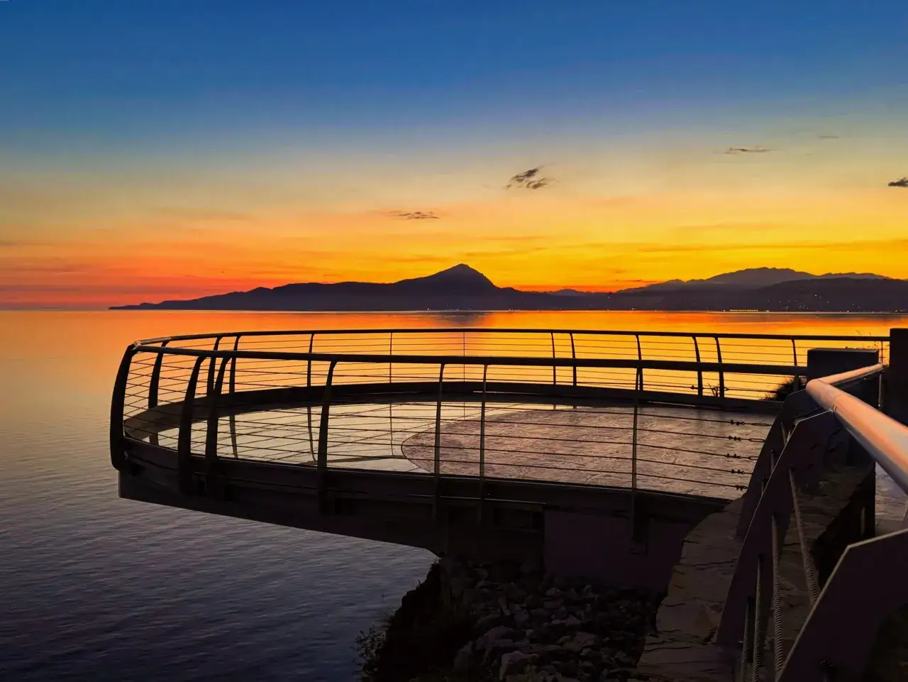Tramonto infuocato sul mare, con la silhouette di una montagna sullo sfondo. Una passerella moderna si protende sull'acqua, offrendo una vista mozzafiato del lungomare di Maratea.