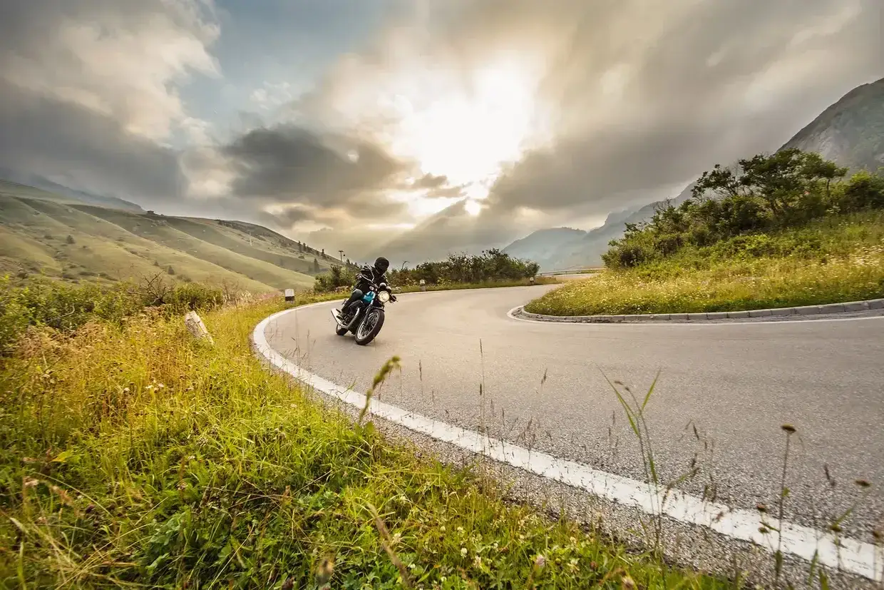 Motociclista in piega su una strada tortuosa al Passo della Contessa, con montagne verdi e cielo nuvoloso.
