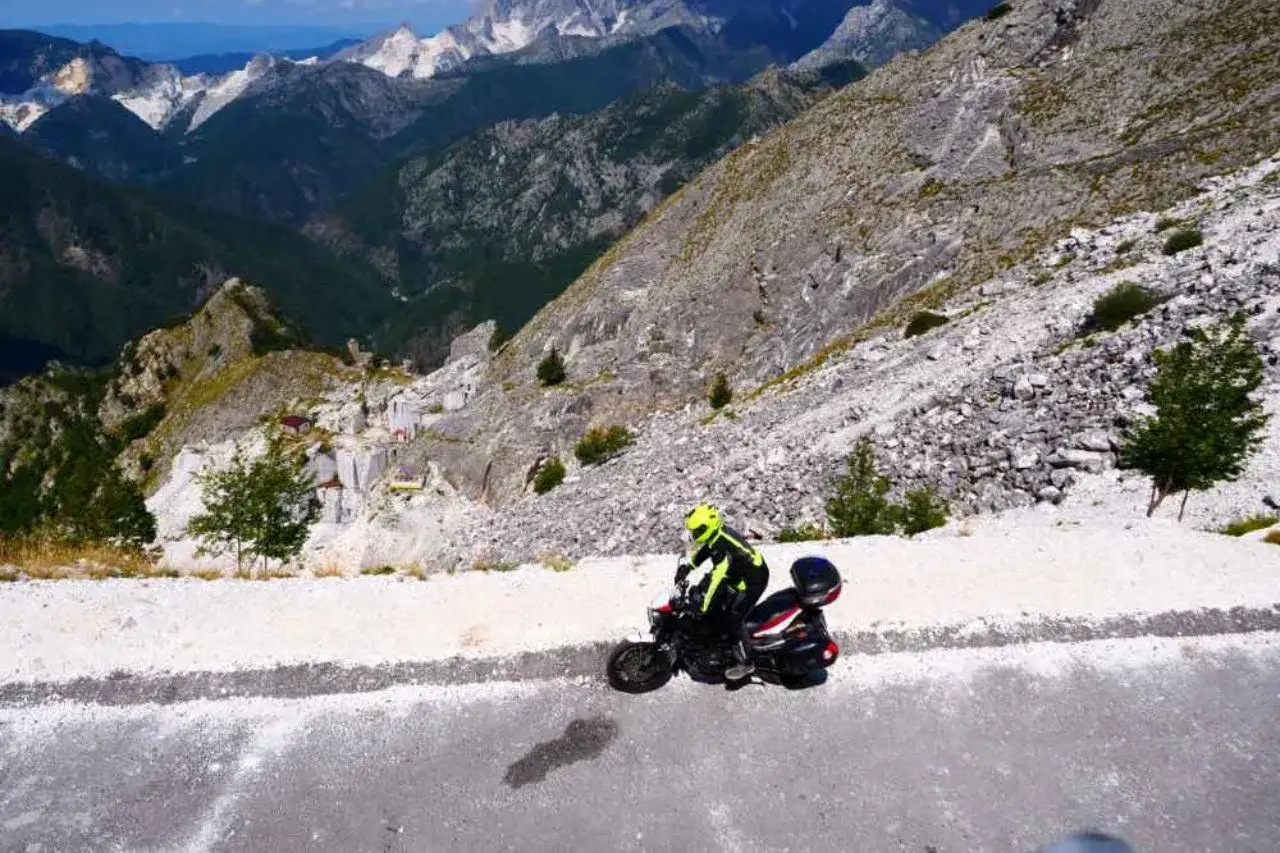 Un motociclista con casco giallo affronta un passo del vestito in moto, tra montagne rocciose e cave di marmo.