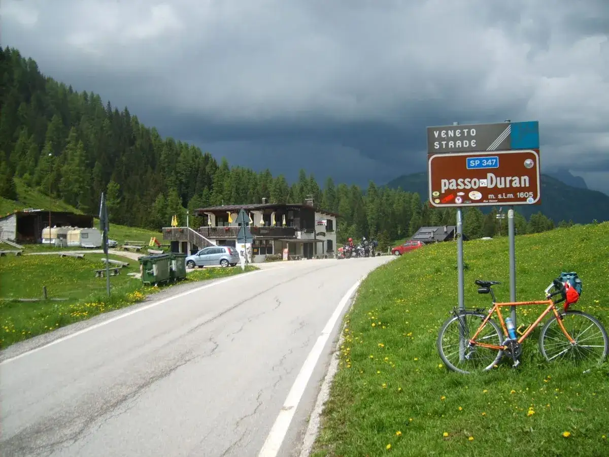 Strada alpina con cartello per passo Duran. Bicicletta arancione parcheggiata su prato fiorito, con sfondo di boschi e cielo minaccioso.