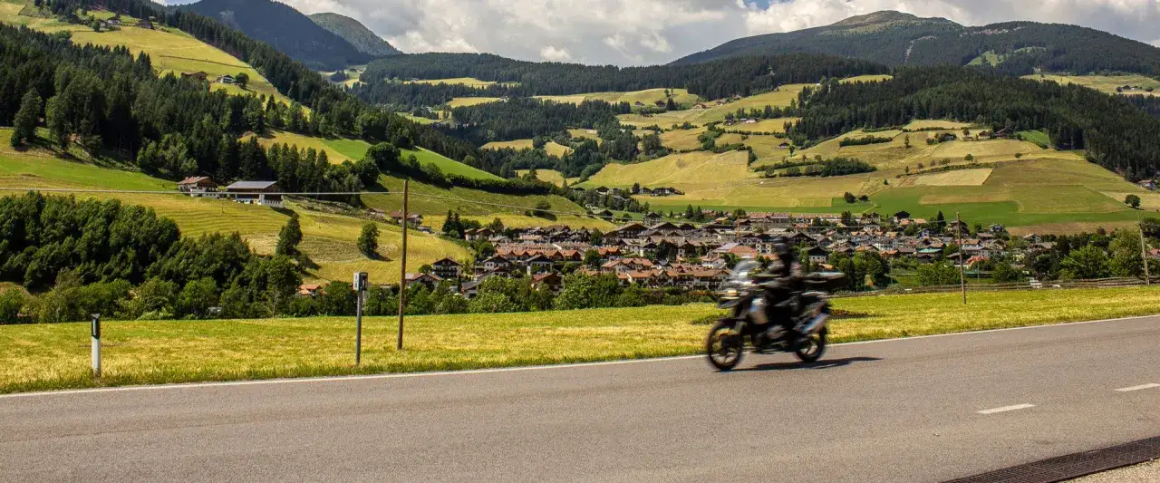 Un motociclista sfreccia lungo una strada panoramica verso un villaggio alpino a Merano, Trentino, circondato da verdi colline e montagne boscose.