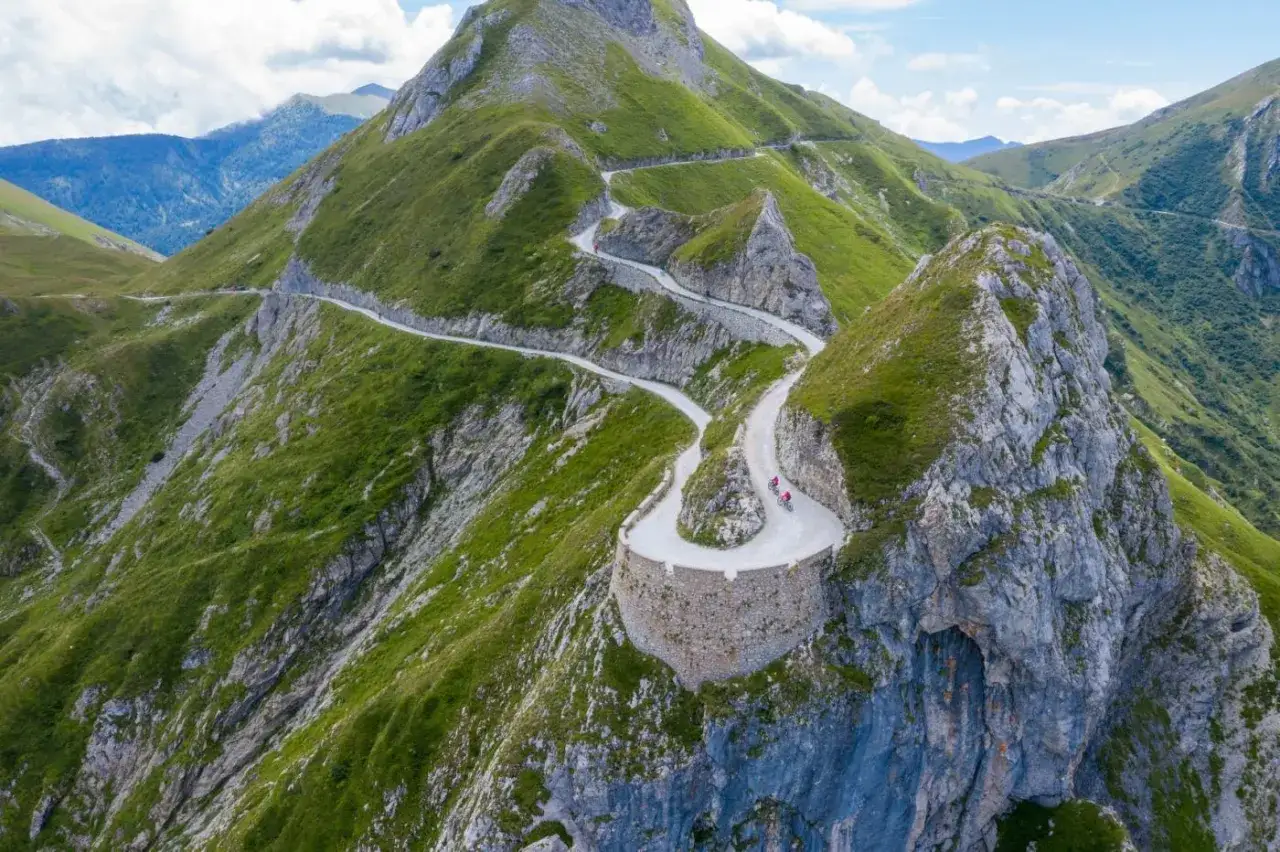 Strada tortuosa con tornante della boaria, due ciclisti affrontano la salita su un passo montano verdeggiante.