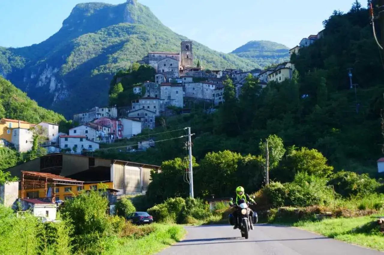 Un motociclista esplora la Lunigiana, con un borgo arroccato su una collina verdeggiante sullo sfondo.