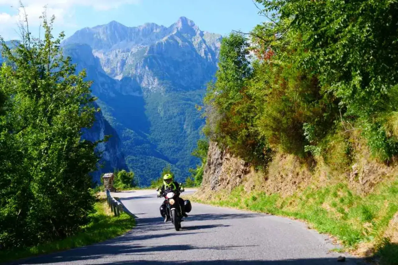 Motociclista in giallo percorre una strada panoramica verso il passo dei Carpinelli, con imponenti montagne sullo sfondo.