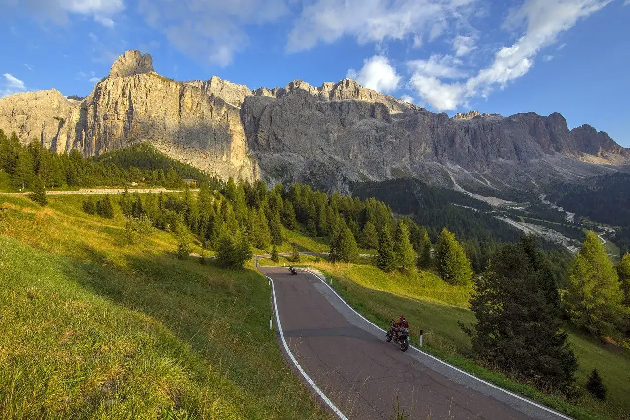 Strada tortuosa tra prati verdi e boschi di conifere, con motociclisti che esplorano le maestose Dolomiti sotto un cielo azzurro e nuvoloso.