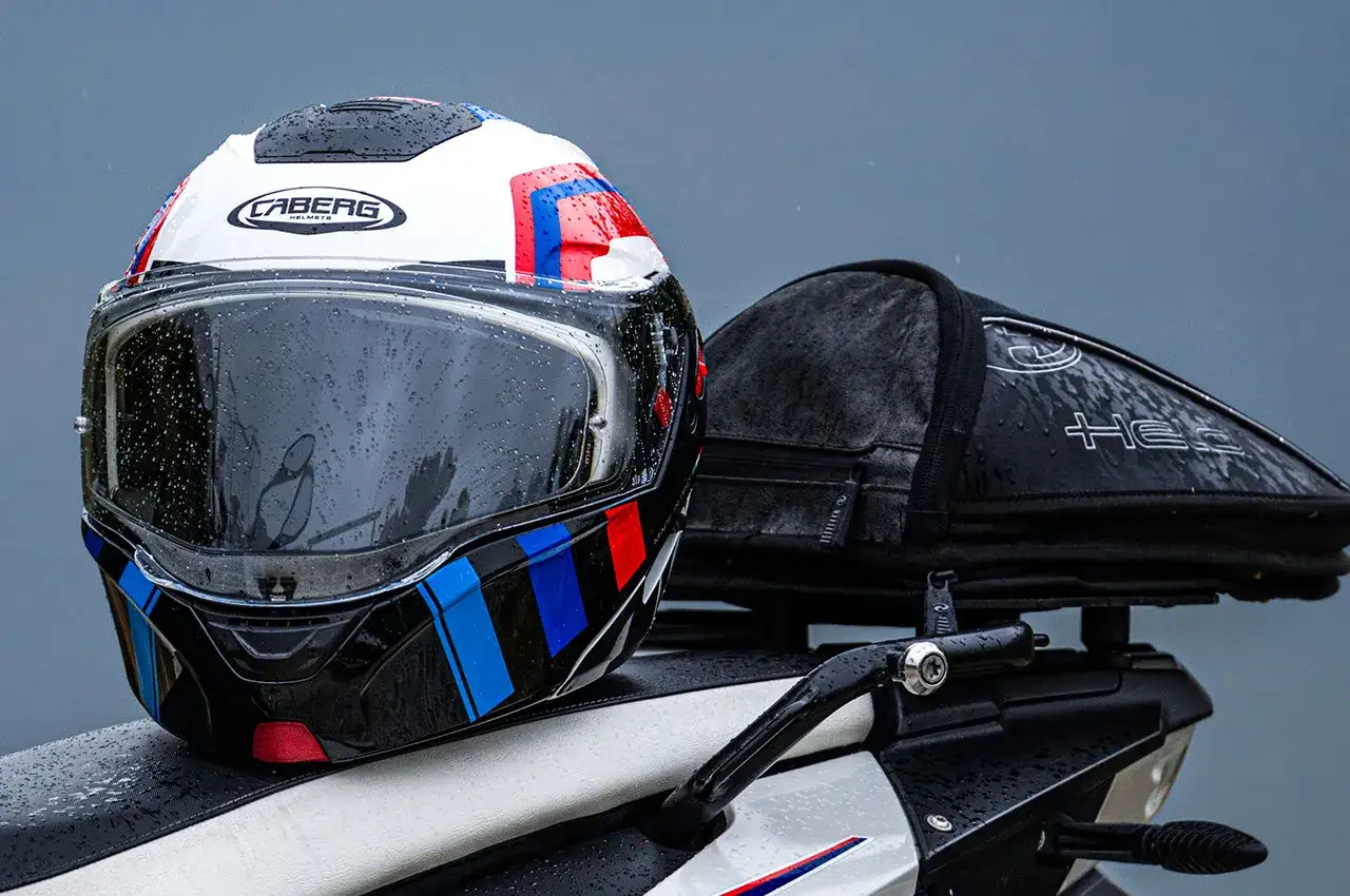 Casco Caberg con visiera priva di graffi, gocce di pioggia sulla superficie. Sullo sfondo, una borsa da moto.
