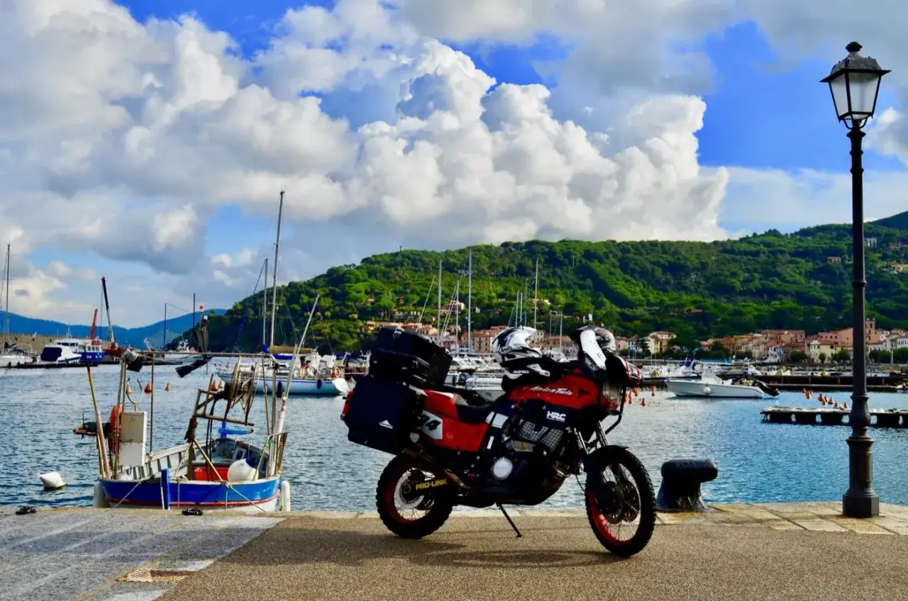 Moto rossa parcheggiata sul molo di Porto Azzurro, Elba, con barche a vela e colline verdi sullo sfondo.