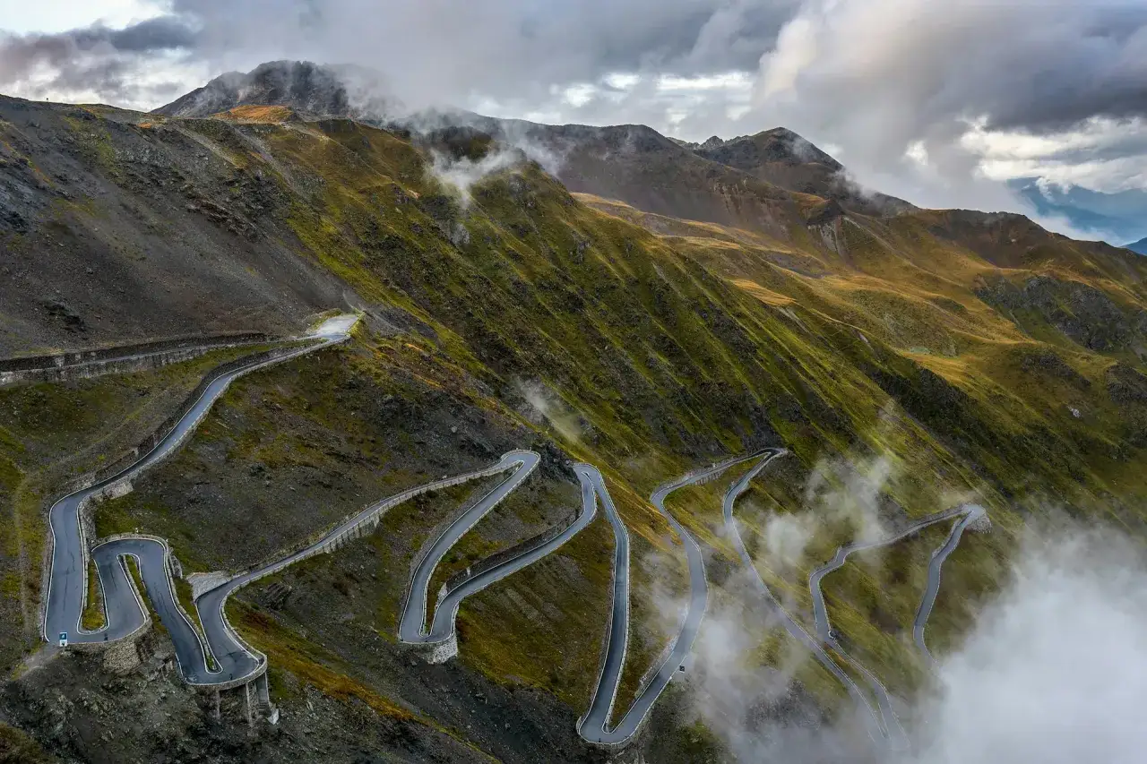 Serpeggiante strada di montagna al passo della forcella, avvolta da nuvole e nebbia, con pendii erbosi e rocciosi.