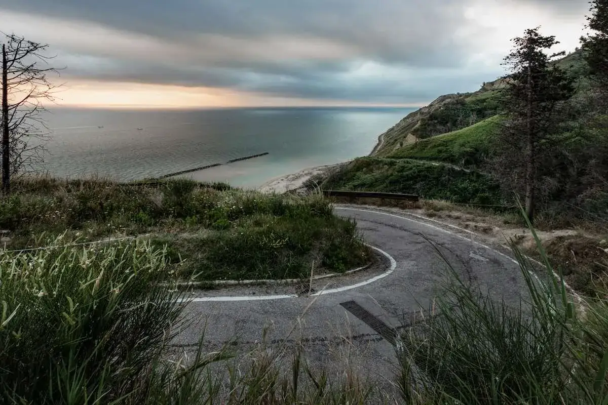 Strada tortuosa con vista sul mare Adriatico e sulla costa verdeggiante. Panoramica Gabicce al tramonto, con nuvole che si diradano.