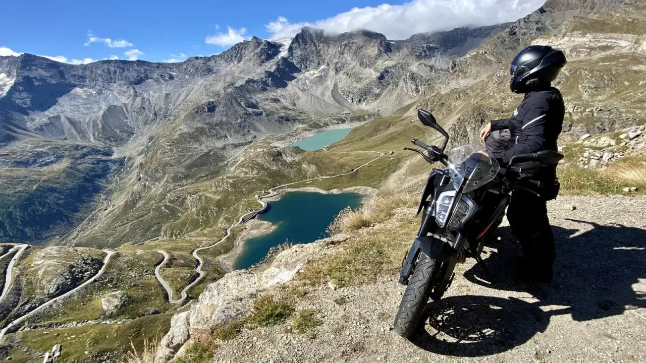 Motociclista con moto ammira il panorama mozzafiato del passo Nivolet, con laghi alpini e tornanti sinuosi.