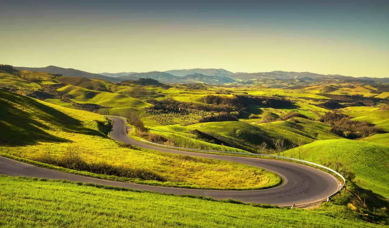 Una sinuosa volterrana strada attraversa dolci colline verdi sotto un cielo limpido.