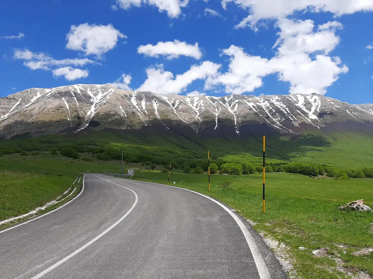 Strada panoramica verso il Passo San Leonardo, con montagne innevate e verdi colline.