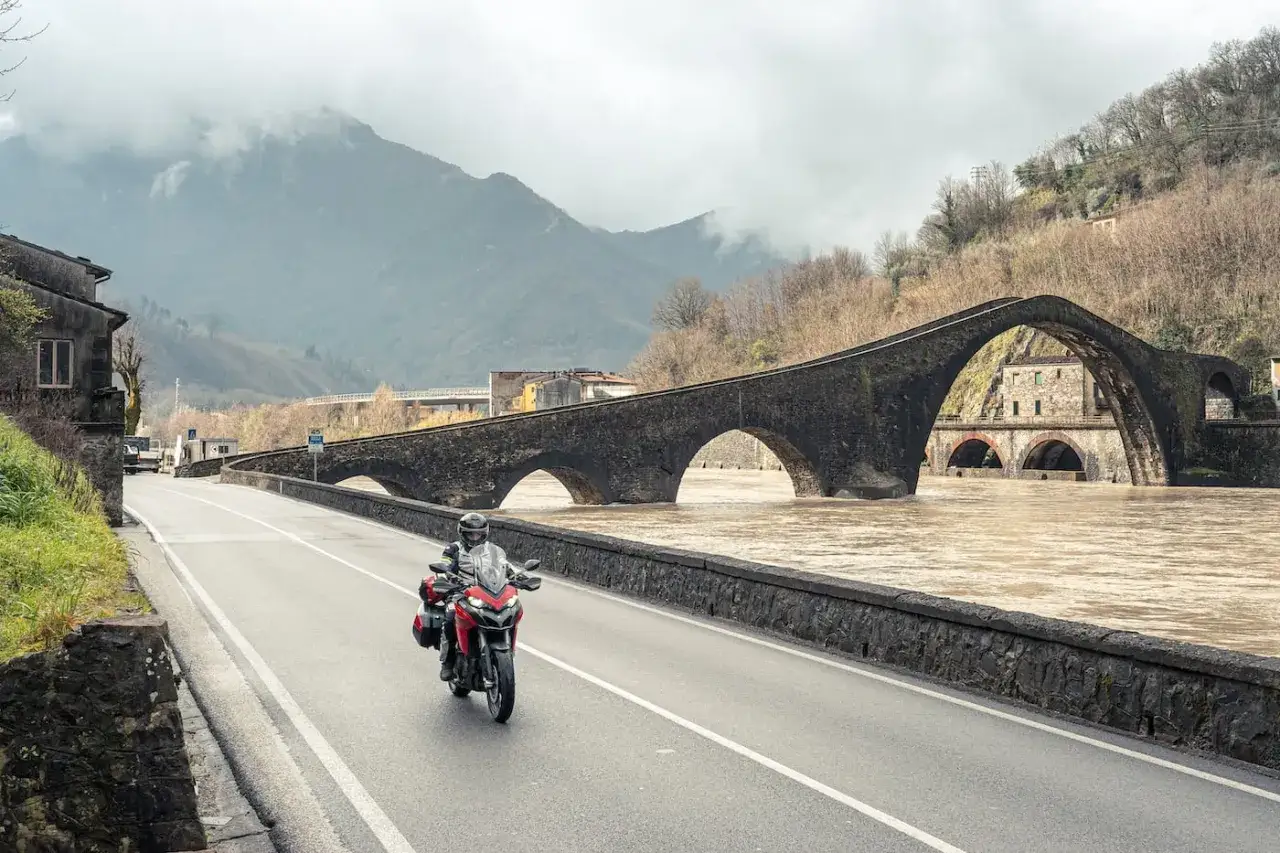 Un motociclista esplora le strade pi&ugrave; belle della Toscana, passando sul Ponte del Diavolo con le montagne nebbiose sullo sfondo.