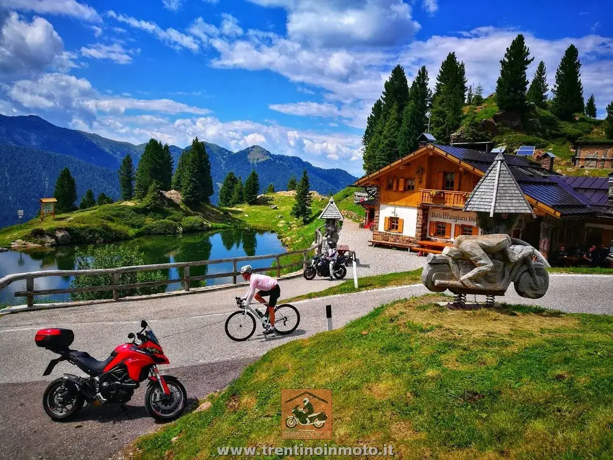Ciclista e moto al Passo Manghen, con rifugio alpino e lago sullo sfondo. Paesaggio montano incantevole.